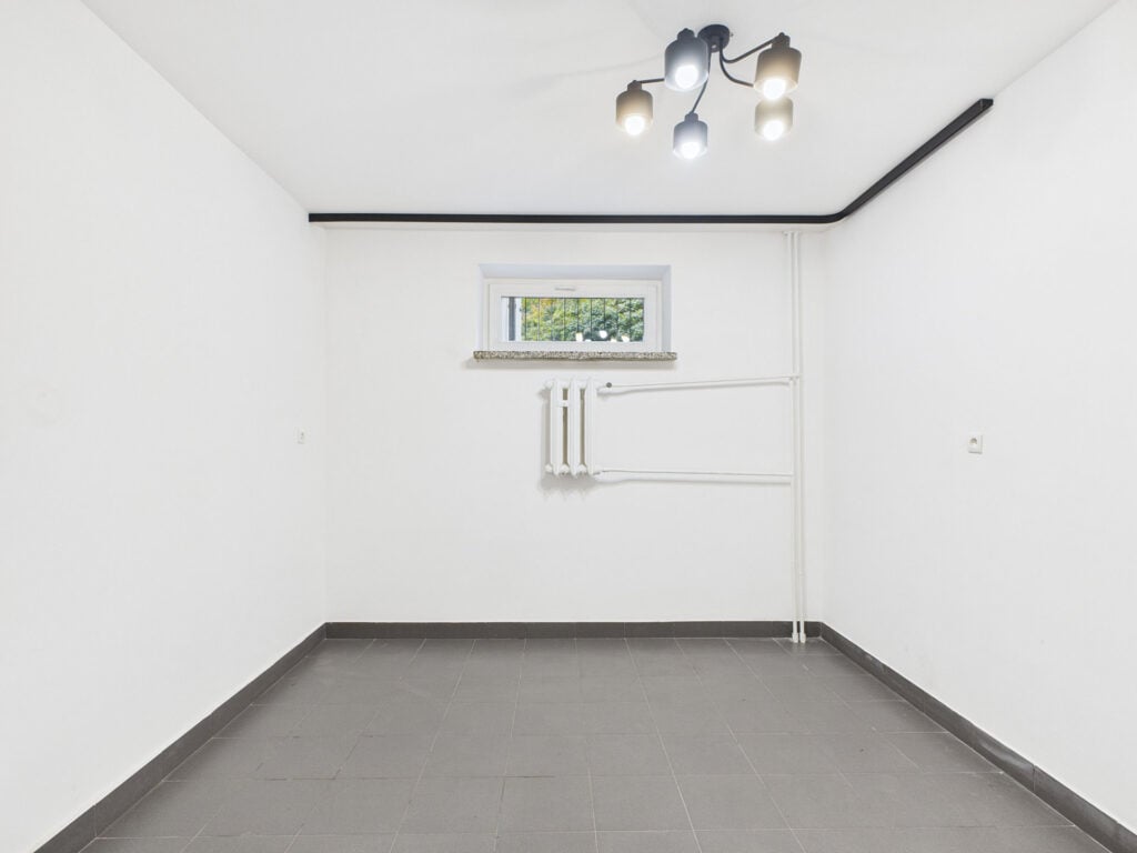 Pusta biała piwnica z oknem i lampą sufitową.