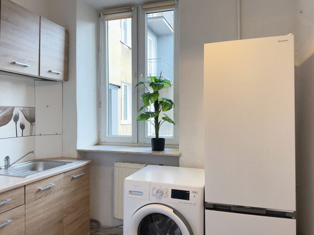 Kuchnia z lodówką, pralką i rośliną na parapecie.